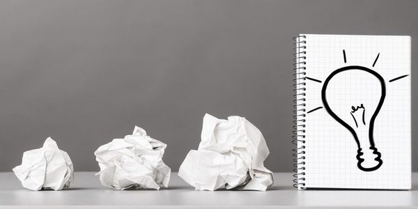 Tres hojas de papel blanco arrugadas alineadas sobre una superficie plana, expresando errores o ideas fallidas, seguidas de una libreta con espiral abierta que muestra un dibujo de una bombilla en papel cuadriculado. La bombilla está rodeada de líneas que simbolizan una idea brillante. El fondo gris resalta los objetos en primer plano, transmitiendo visualmente el proceso creativo: desde ideas descartadas hasta una inspiración exitosa.