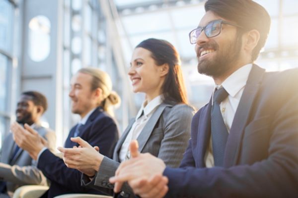 Cuatro personas vestidas con ropa de oficina están sentadas en fila, aplaudiendo con entusiasmo. Parecen estar en un evento profesional o conferencia, en un espacio moderno y bien iluminado con grandes ventanales y estructuras metálicas visibles al fondo. Todas miran en la misma dirección, mostrando atención y aprobación hacia lo que ocurre fuera del encuadre. La escena transmite un ambiente de profesionalismo, participación activa y éxito del evento.