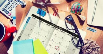 Mesa de trabajo con un calendario abierto en el centro, marcado el día 17 con un círculo rojo. Alrededor hay gafas, notas adhesivas de colores, lápices, clips, una taza de café, una calculadora, un bloc de notas, una pelota de gomas elásticas y papeles arrugados, transmitiendo la idea de planificación y organización de eventos.